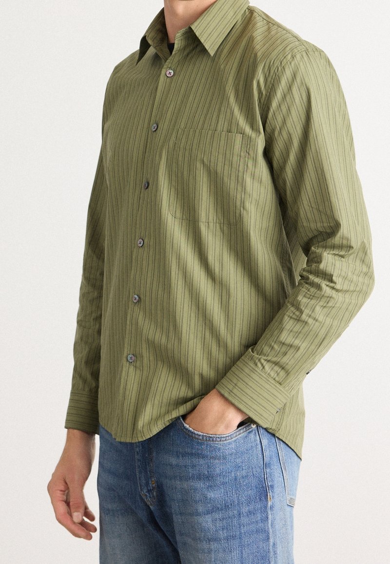 Homme portant une chemise olive à manches longues boutonnée avec de fines rayures verticales et un jean bleu, une main dans la poche.