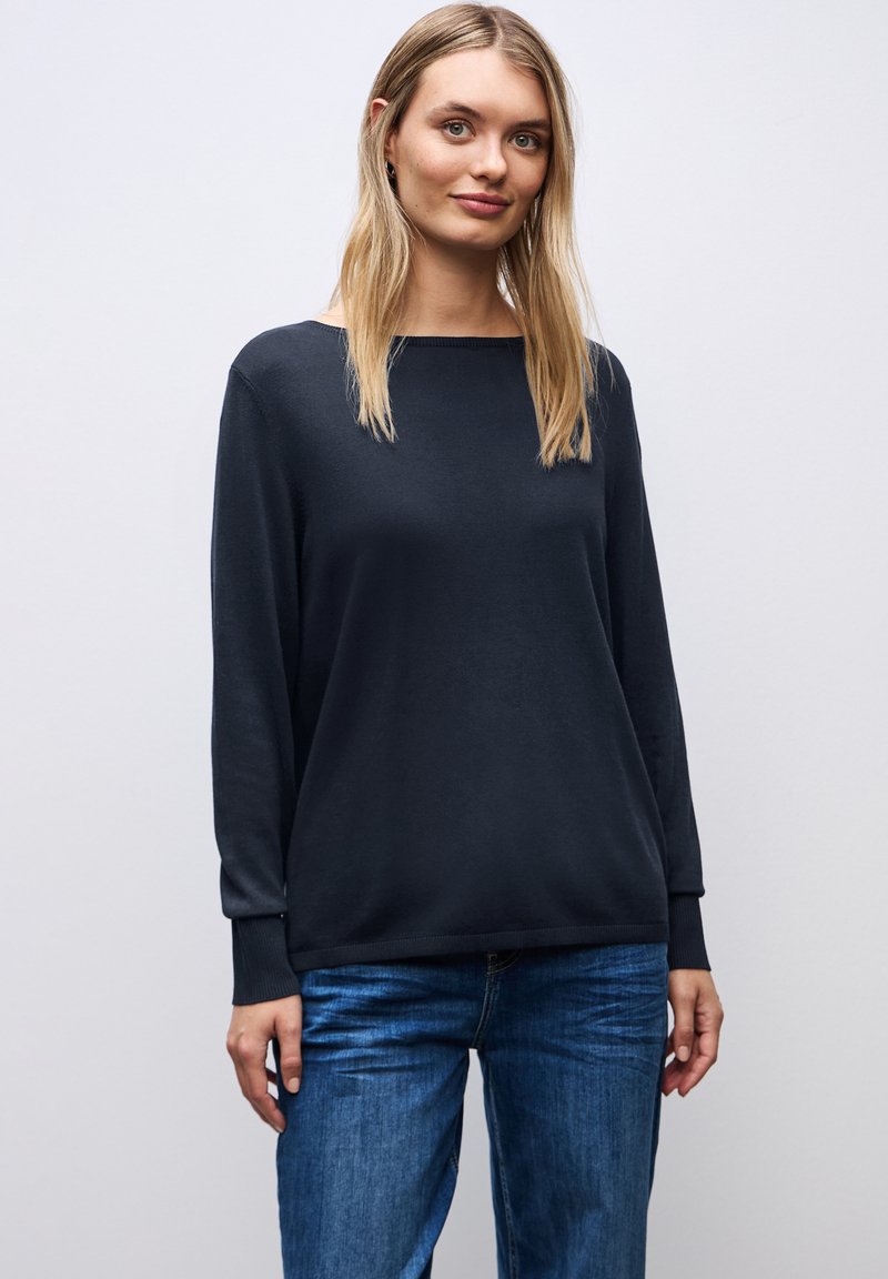 Schwarzer Pullover mit langen Ärmeln aus einem weichen Material, mit einem breiten Ausschnitt und gerippten Bündchen. Kombiniert mit blauen Jeans steht das Modell vor einem neutralen Hintergrund.