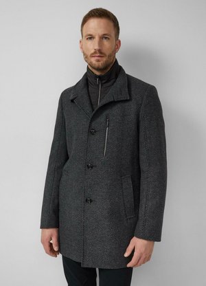 Man draagt een donkergrijze getextureerde jas met een borstzak met rits over een zwarte binnenlaag met rits, staand tegen een effen lichtgrijze muur.