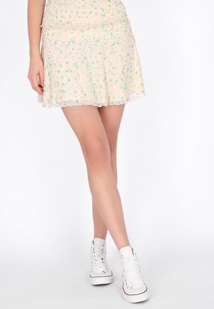 Kobieta ubrana w lekką, kwiecistą mini spódniczkę i białe wysokie trampki, stojąca ze skrzyżowanymi nogami na jednolitym białym tle.