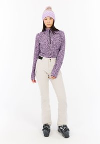 Sudadera morada con estampado de leopardo y cierre en la parte superior, combinada con pantalones de esquí beige y botas de esquí negras con acentos multicolores. El gorro con pompom es lavanda.