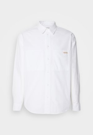 White long-sleeve button-up shirt made of cotton. Features two chest pockets, a curved hem, and a small gold logo on the left pocket.