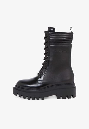 Bottine en cuir noir avec une semelle plateforme épaisse, bout rond, accents côtelés et lacets passant par des œillets métalliques. Fini hautement poli.