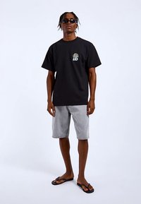 Svart bomulls-T-shirt med logotyp på bröstet, kombinerad med ljusgrå denimshorts och svarta flip-flops. Modellen står mot en vit bakgrund.