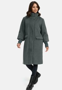 Manteau en polaire vert avec un col haut, fermeture à l'avant, deux poches et poignets ajustés, associé à des bottines noires et un jean foncé.