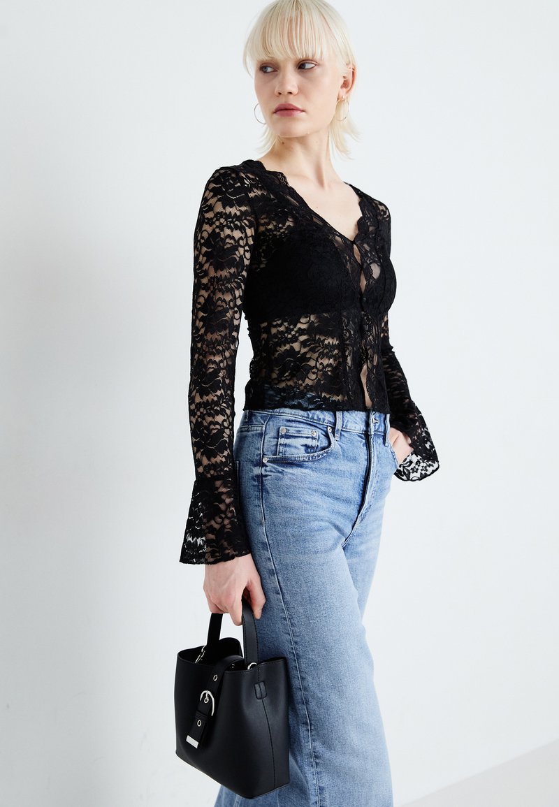 Top en dentelle noire à manches longues avec des motifs floraux, décolleté en V et manches évasées, associé à un jean en denim bleu clair et un petit sac à main noir.