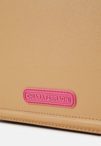 Hnědý kožený doplněk s texturovaným povrchem a růžovou embosovanou nálepkou s logem. Obsahuje šité detaily pro zvýšení odolnosti.