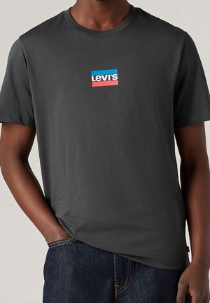 Mężczyzna w ciemnoszarej koszulce Levi's z małym niebiesko-czerwonym logo na piersi oraz ciemnoniebieskich dżinsach, z ręką w kieszeni.