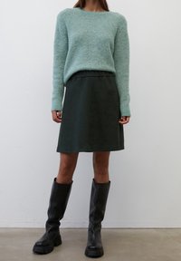 Suéter esponjoso verde claro con mangas largas, combinado con una falda de cinturilla elástica verde oscuro y botas negras de caña alta y gruesa.