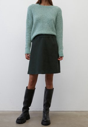 Maglione fuzzy verde chiaro con maniche lunghe, abbinato a una gonna con cintura elastica verde scuro e stivali neri alti fino al ginocchio con tacco grosso.