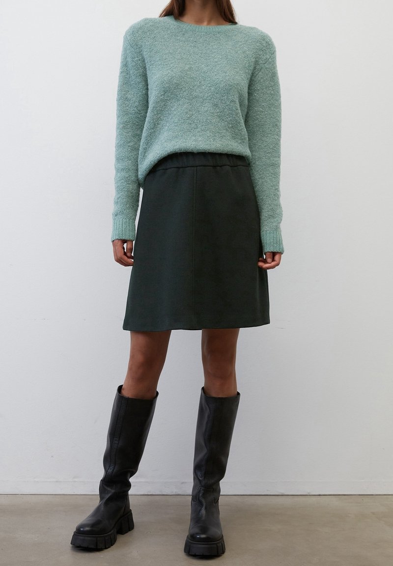 Suéter esponjoso verde claro con mangas largas, combinado con una falda de cinturilla elástica verde oscuro y botas negras de caña alta y gruesa.