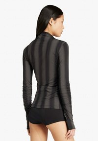 Donna con capelli lunghi e scuri che indossa un top nero aderente a maniche lunghe con righe verticali e pantaloncini neri, rivolta verso destra su uno sfondo bianco.