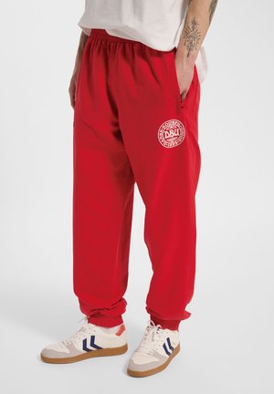 Osoba nosząca czerwone spodnie dresowe z logo Duńskiego Związku Piłki Nożnej, białe buty sportowe z grafiką oraz białą koszulę wpuszczoną w talię.