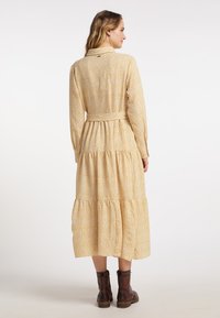 Langsleeve, gele bloemenjurk met een gelaagde rok, een ceintuur en kraagdetails. Gstyled met bruine enkellaarsjes. Zachte stoftextuur.