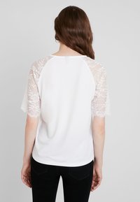 Vit topp med spetskortärmar. Mjukt material, rundad halsringning och raglanärmsdesign. Kullrig rygg med lätt kurva vid fållen.