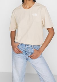 T-shirt in cotone beige con scollo rotondo, maniche corte e logo bianco sul lato sinistro del petto; indossato con jeans a vita alta di colore blu chiaro.