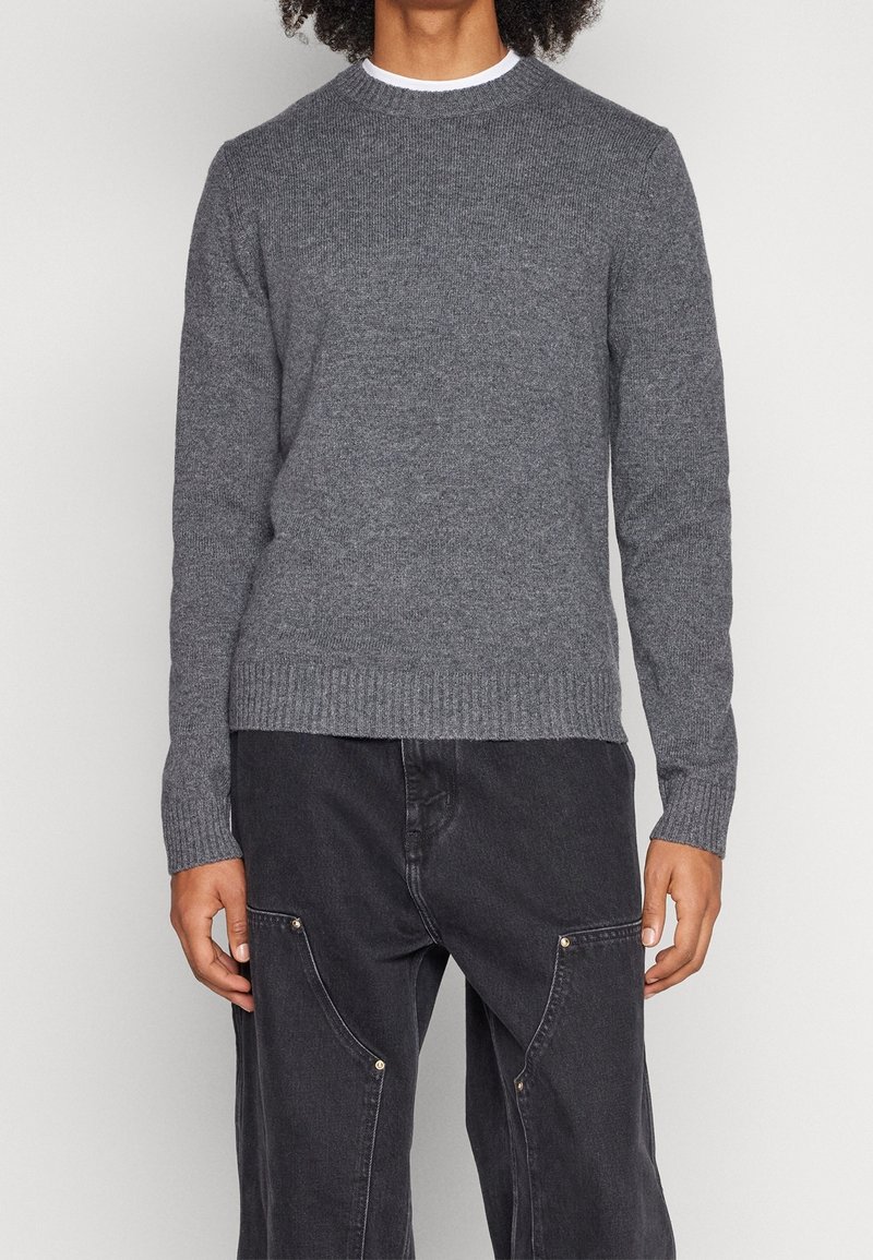 Personne portant un pull en maille gris cintré et un jean noir ample avec des poches avant, debout devant un arrière-plan clair uni.