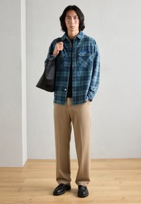 Blå og grønn rutete skjorte med knapper, beige bukser, svarte sko og en svart tote-bag. Glatte teksturer og avslappet silhuett.