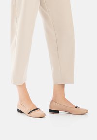 Des chaussures plates beiges avec un bout carré, un détail de sangle noire à l'avant et des talons bas, associées à un pantalon beige coupé.