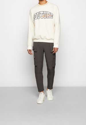 Beżowa bluza z napisem "COUTURE", w połączeniu z ciemnoszarymi spodniami typu cargo z kieszeniami bocznymi. Stylizowana z białymi trampkami.