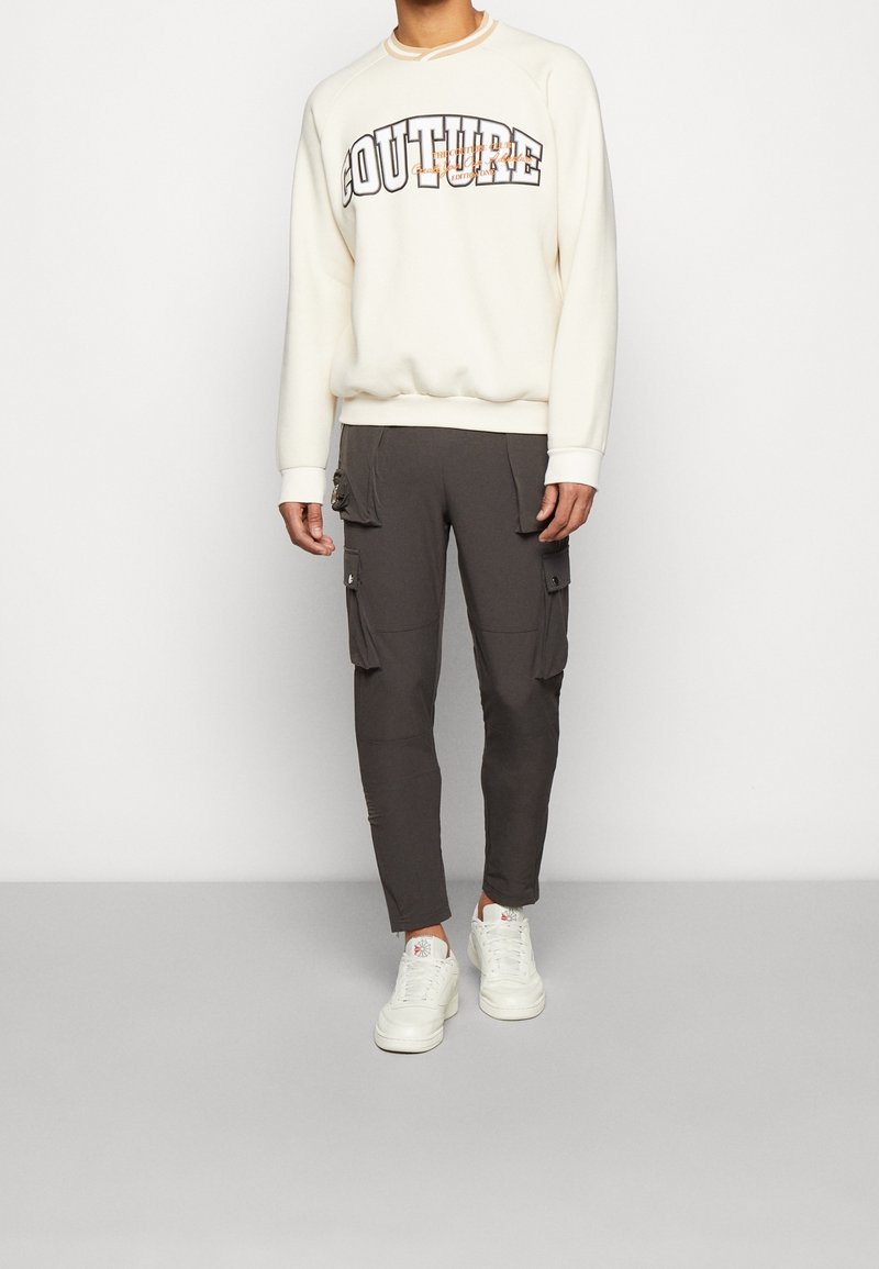 Beige sweatshirt with "COUTURE" graphic, paired with dark gray cargo pants featuring side pockets. Styled with white sneakers.