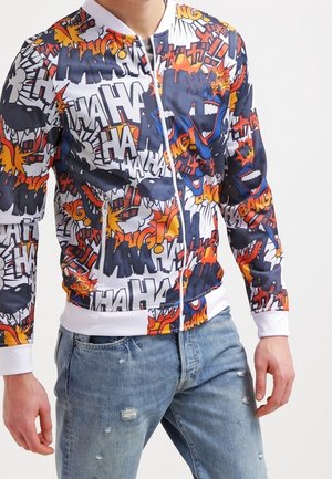 Man met een kleurrijke ritssluiting jas met stripstijl "HA," "BANG," en explosiegrafieken, gecombineerd met gescheurde blauwe jeans.