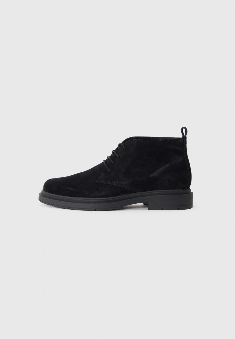 Botas de tobillo negras de ante con una textura suave, diseño con cordones y suela de goma negra. Cuenta con una lengüeta en la parte trasera para facilitar su uso.