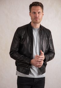 Black leather jacket with a zip front, stand collar, and ribbed cuffs. Worn over a gray t-shirt and paired with black jeans.