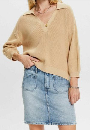 Kobieta w beżowym dzianinowym swetrze na jeden guzik i jasnej, niebieskiej spódnicy dżinsowej z przednimi kieszeniami, ręka na biodrze, białe tło.