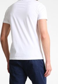 Man wearing a plain white short-sleeve t-shirt and dark blue pants, seen from the back against a light background.