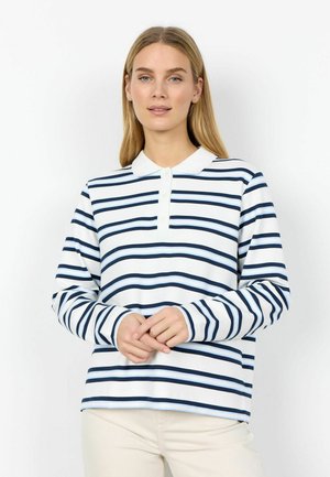 Kvinde med langt blondt hår iført en hvid polo-skjorte med marineblå og lyseblå vandrette striber, stående med hænderne foldet.