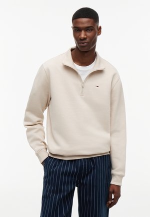 Muškarac u bež puloveru s četvrtastim patentnim zatvaračem preko bijele košulje, uparenom s tamnoplavam hlačama s tankim prugama, stoji sa rukama u džepovima.