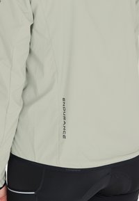 Lys grå vindjakke med en glat tekstur; har et "ENDURANCE" logo i sort på siden; parret med sorte bukser.