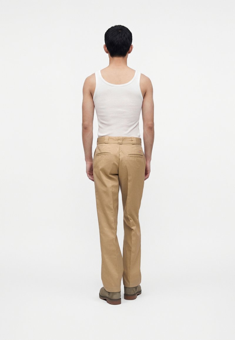 Uomo con capelli corti e neri che indossa una canottiera bianca, pantaloni beige e scarpe marroni, in piedi di spalle davanti a uno sfondo bianco semplice.