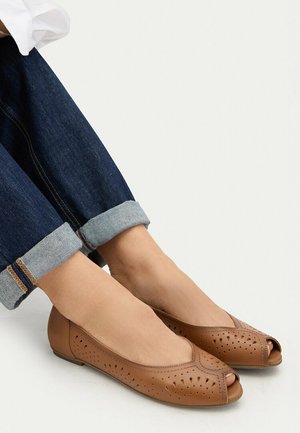 Pies calzando zapatos planos de cuero perforado color beige con punta abierta, combinados con jeans oscuros con puños, sobre un fondo blanco liso.