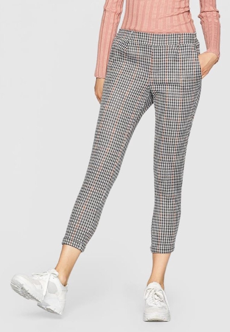 Pantalon slim fit longueur cheville avec un motif à carreaux noir et blanc, porté avec des baskets blanches et un top rose côtelé à manches longues.