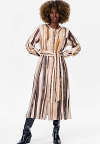 Lange, knoopsluiting jurk in gestreept beige, bruin en gouden patroon. Beschikt over een riem in de taille, lange mouwen en een vloeiend, lichtgewicht stof.