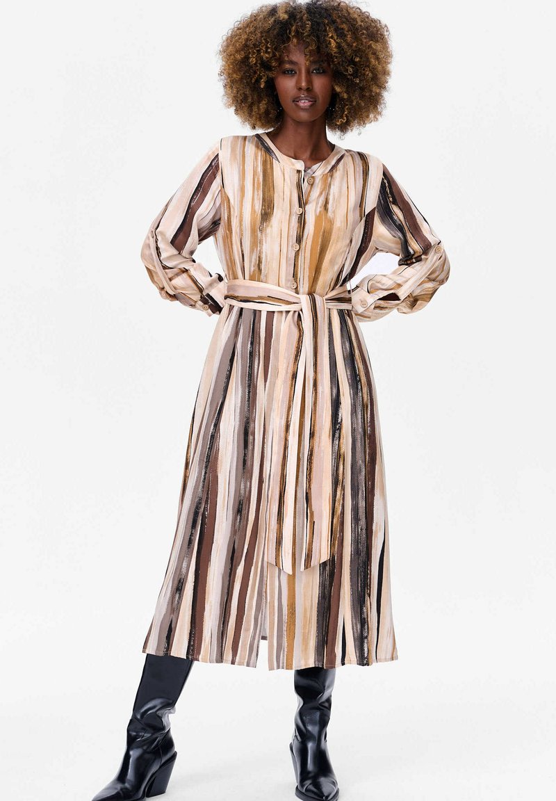 Lange, knoopsluiting jurk in gestreept beige, bruin en gouden patroon. Beschikt over een riem in de taille, lange mouwen en een vloeiend, lichtgewicht stof.