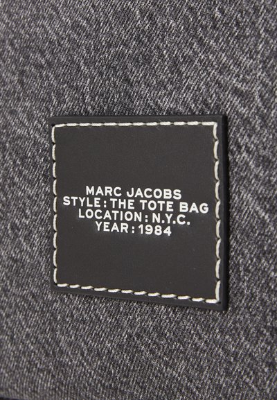 Sac fourre-tout en denim gris avec une étiquette en cuir noir. L'étiquette présente un texte blanc indiquant la marque et les détails : "MARC JACOBS," "LE SAC FOURRE-TOUT," "N.Y.C.," "1984."