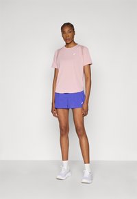 Ροζ ανοιχτό ροζ κοντομάνικο πουκάμισο με λογότυπο, συνδυασμένο με μπλε αθλητικό σορτς. Το μοντέλο φοράει λευκά αθλητικά παπούτσια και κάλτσες μέχρι τον αστράγαλο, στέκεται μπροστά σε ουδέτερο φόντο.