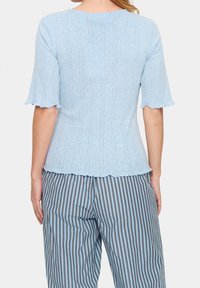 Ljusblå topp med korta, utsvängda ärmar. Har en ribbad textur och vågig nederkant. Matchas med grå och blå randiga byxor.