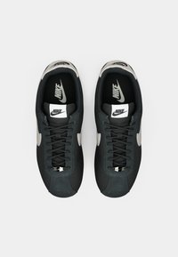 Nike sko med en sort overdel, grå accenter og en tekstureret tåhætte. Har flade snørebånd og en polstret krave med et logo på tungen.