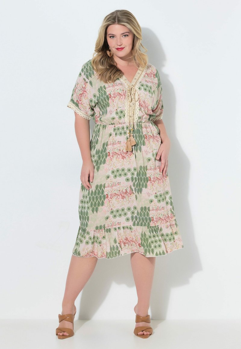 Mujer de cabello rubio viste un vestido de longitud media con estampado floral verde y rosa, cuello con lazo de borlas y sandalias de tacón color canela.