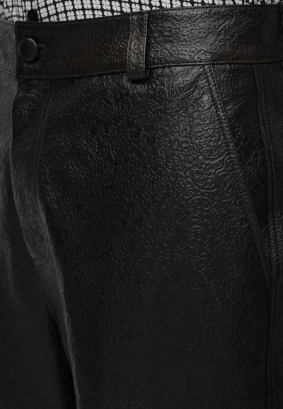 Close-up de calças de couro preto texturizado com padrões florais em relevo e um passador de cinto de couro sobre uma cintura de tecido xadrez.