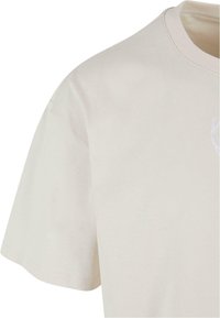Lys beige t-skjorte med korte ermer, rund hals og subtil tekstur; inkluderer en liten brodert detalj på forsiden.