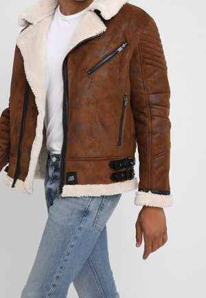 Homme portant une veste en cuir d’agneau marron avec des poches zippées et un jean bleu, marchant sur un fond blanc uni.