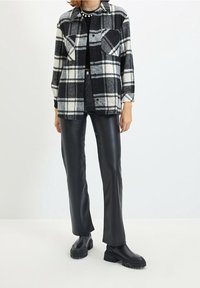 Camicia in flanella a quadri in bianco e nero con due taschini sul petto, abbinata a pantaloni in pelle nera e stivali neri spessi. Tessuto liscio e vestibilità rilassata.