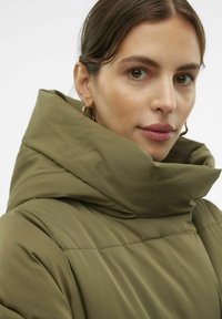Abrigo de pluma verde oliva con un cuello alto y plegado. Textura suave con costuras a lo largo de los hombros. La modelo lleva pendientes de aro dorados.