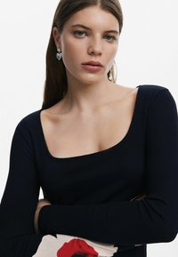 Top lung cu mânecă lungă, negru, cu un decolteu pătrat larg. Modelul poartă cercei argintii lungi. Țesătura texturată și designul ajustat pun în evidență silueta.