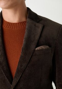 Bordeauxkleurige corduroy blazer met een notch lapel, gecombineerd met een textuurvolle roestkleurige crewneck trui. De zak bevat een gevouwen bruine zakdoek.